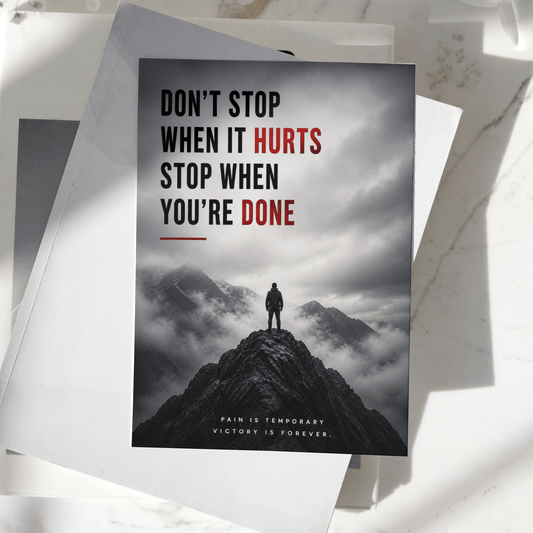 Don't Stop When It Hurts Stop When You're Done motivational poster displayed on a desk, featuring a lone figure standing on a mountain peak, premium inspirational wall art print.