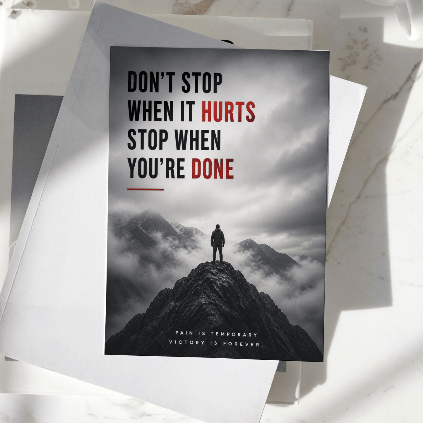 Don't Stop When It Hurts Stop When You're Done motivational poster displayed on a desk, featuring a lone figure standing on a mountain peak, premium inspirational wall art print.